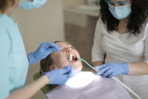 A woman receiving dental treatment while reclined in a dentist's chair, with a dentist and assistant both wearing blue gloves and surgical masks for hygiene. The dentist is examining the patient's teeth with a dental mirror, and an overhead light illuminates the scene for clear visibility.