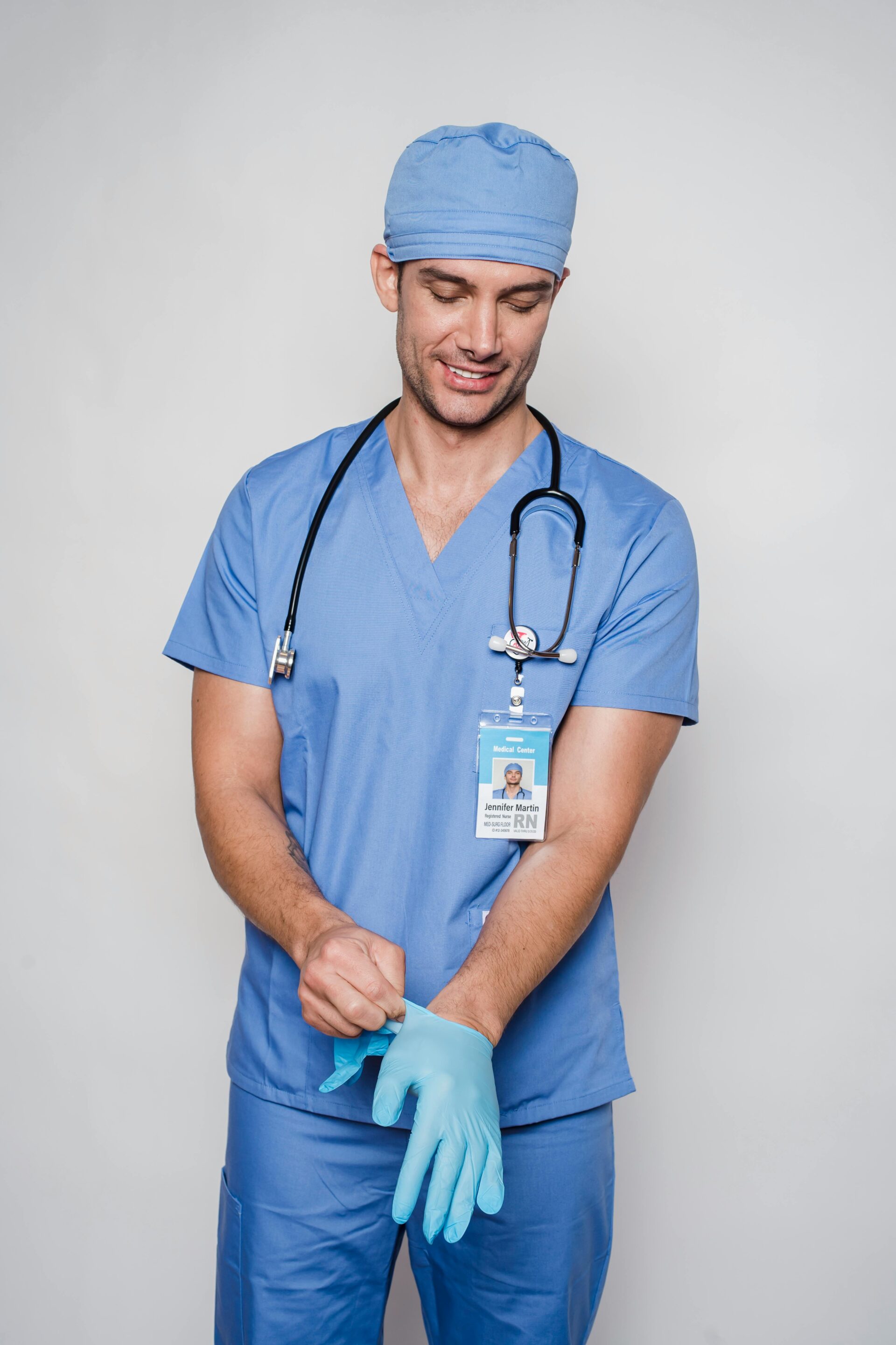 A smiling male nurse in blue scrubs putting on medical gloves, with a stethoscope around his neck and an ID badge reading 'Jennifer Martin, RN'