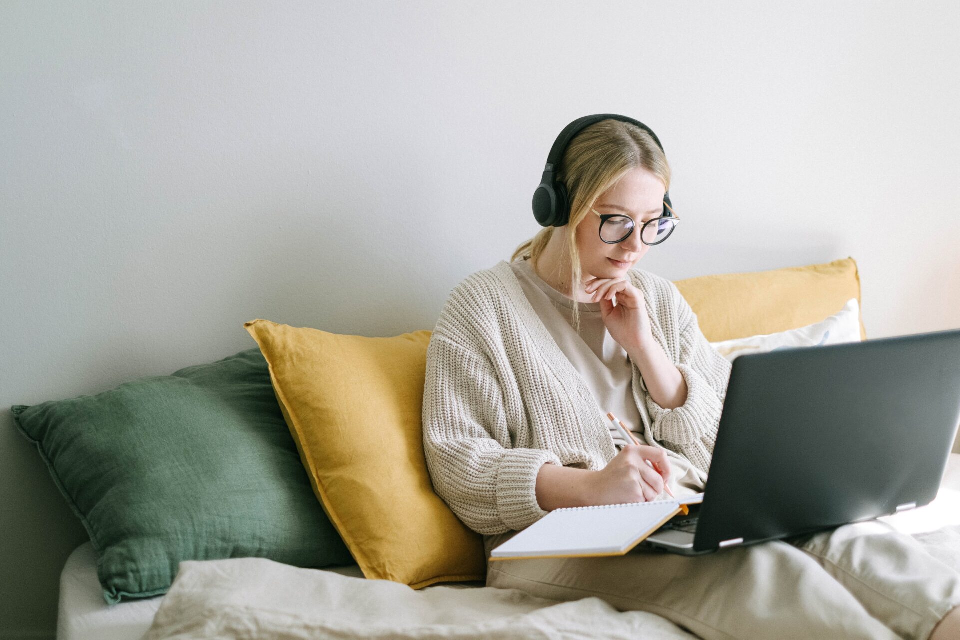 A woman in a beige cardigan and glasses, sitting on a bed with green and yellow cushions, working intently on a laptop while wearing headphones and holding a pen to her chin in thought. She appears to be focused on her task, possibly studying or working from home.