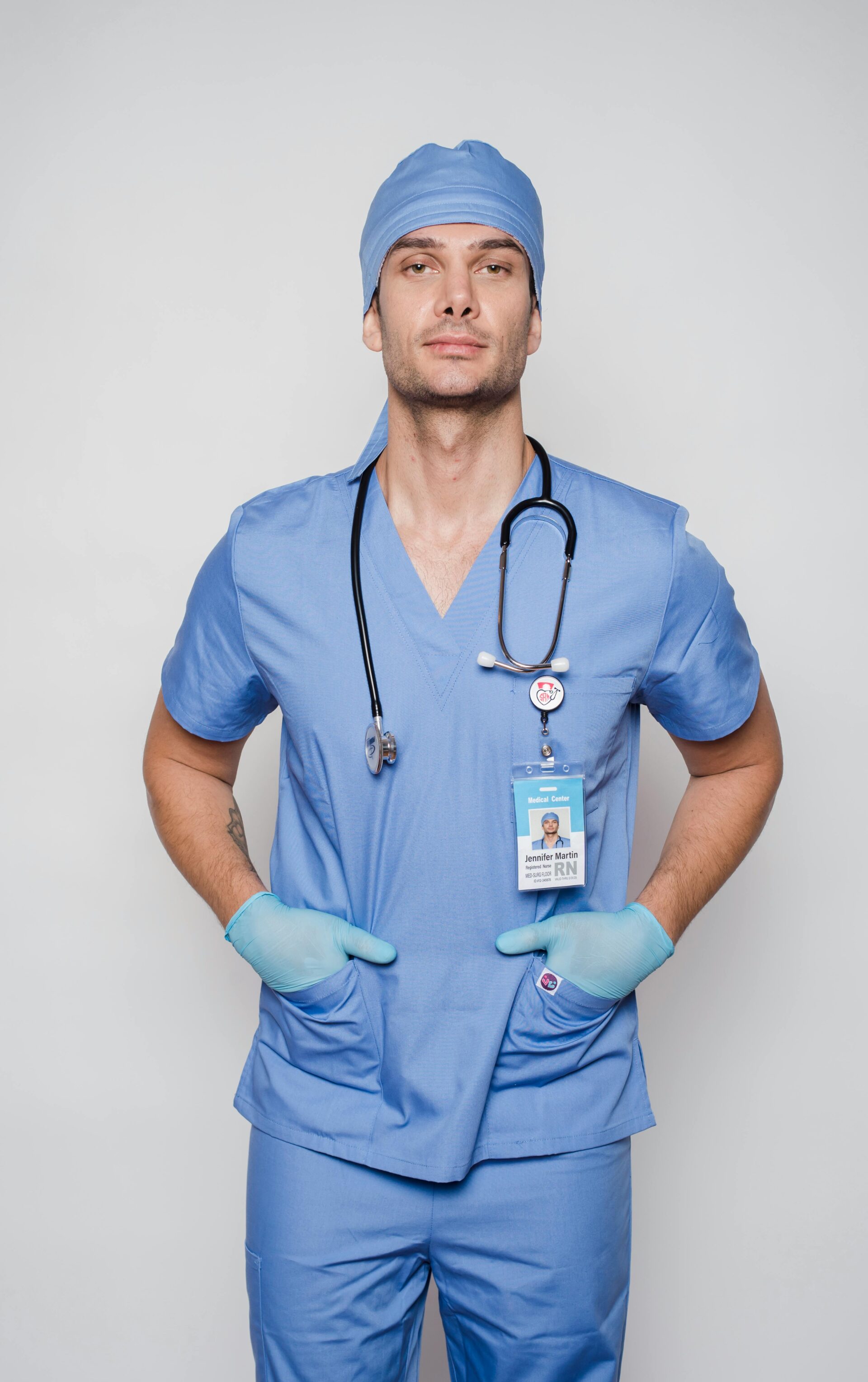 Confident male nurse in blue scrubs with stethoscope and ID badge standing with hands on hips.