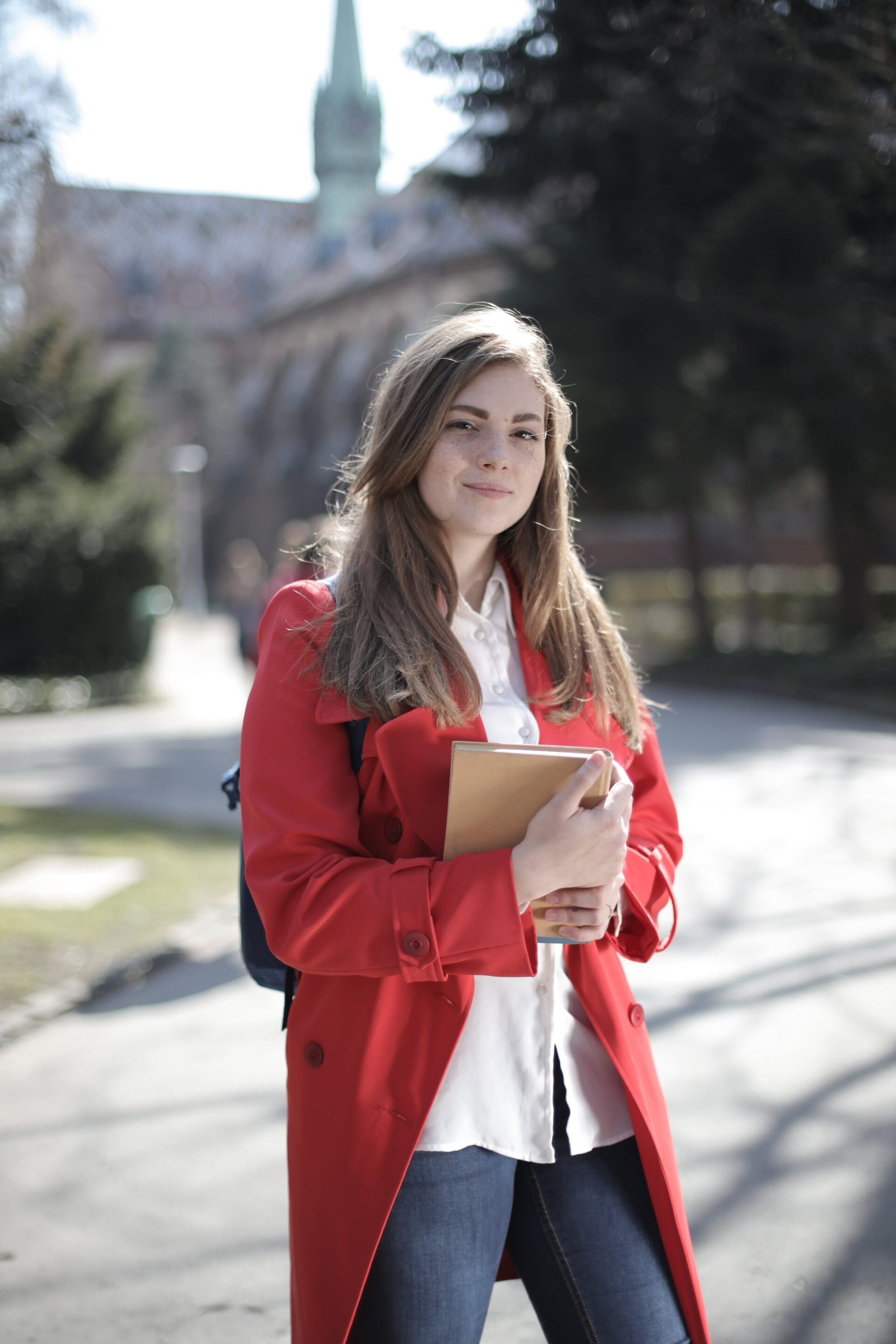 Confident female student in a red coat holding a book on a sunny campus