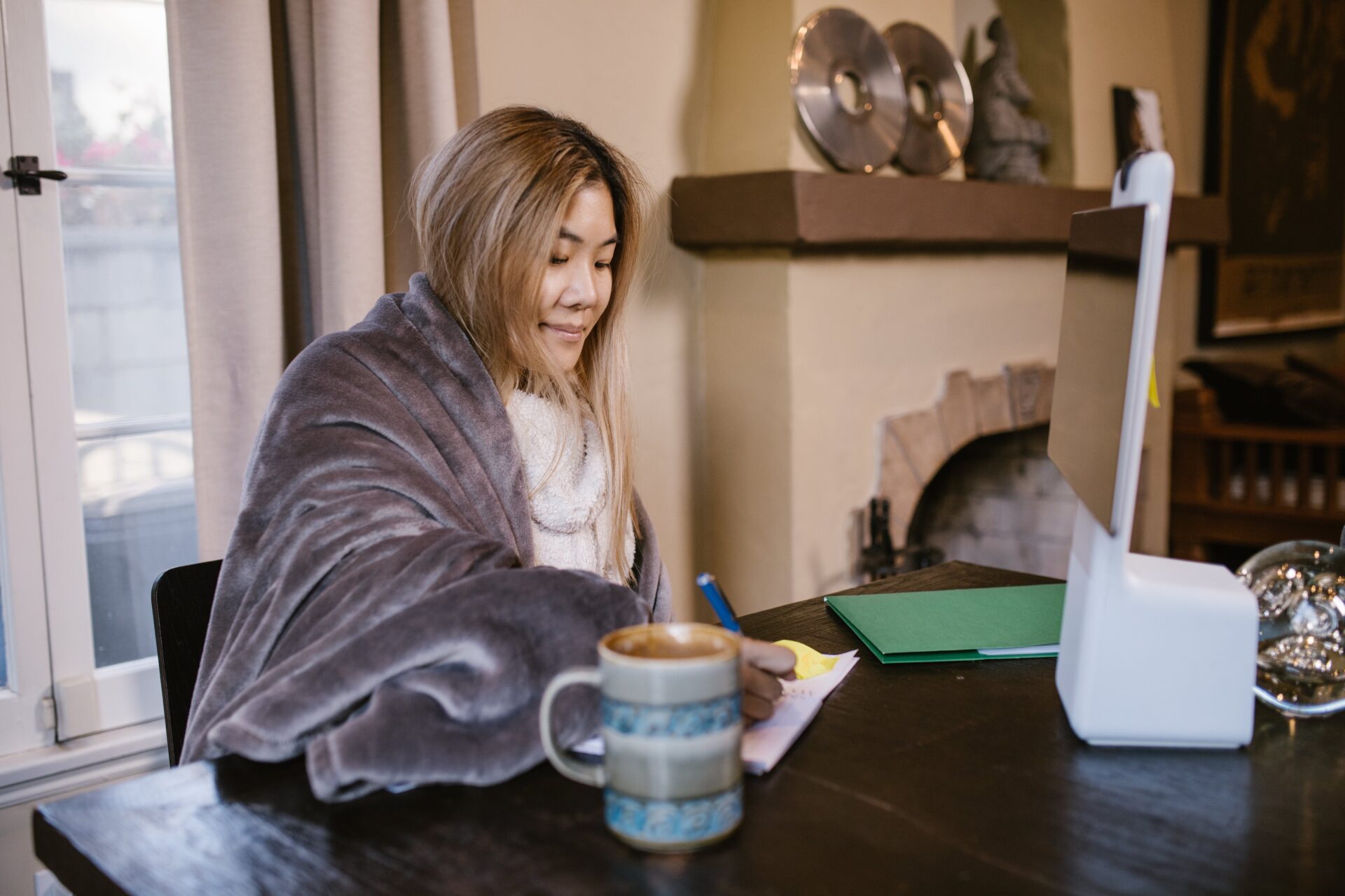 Focused nursing student working on an assignment at home, wrapped in a cozy blanket, with a notepad, pen, and a mug of coffee on the desk, beside an open laptop