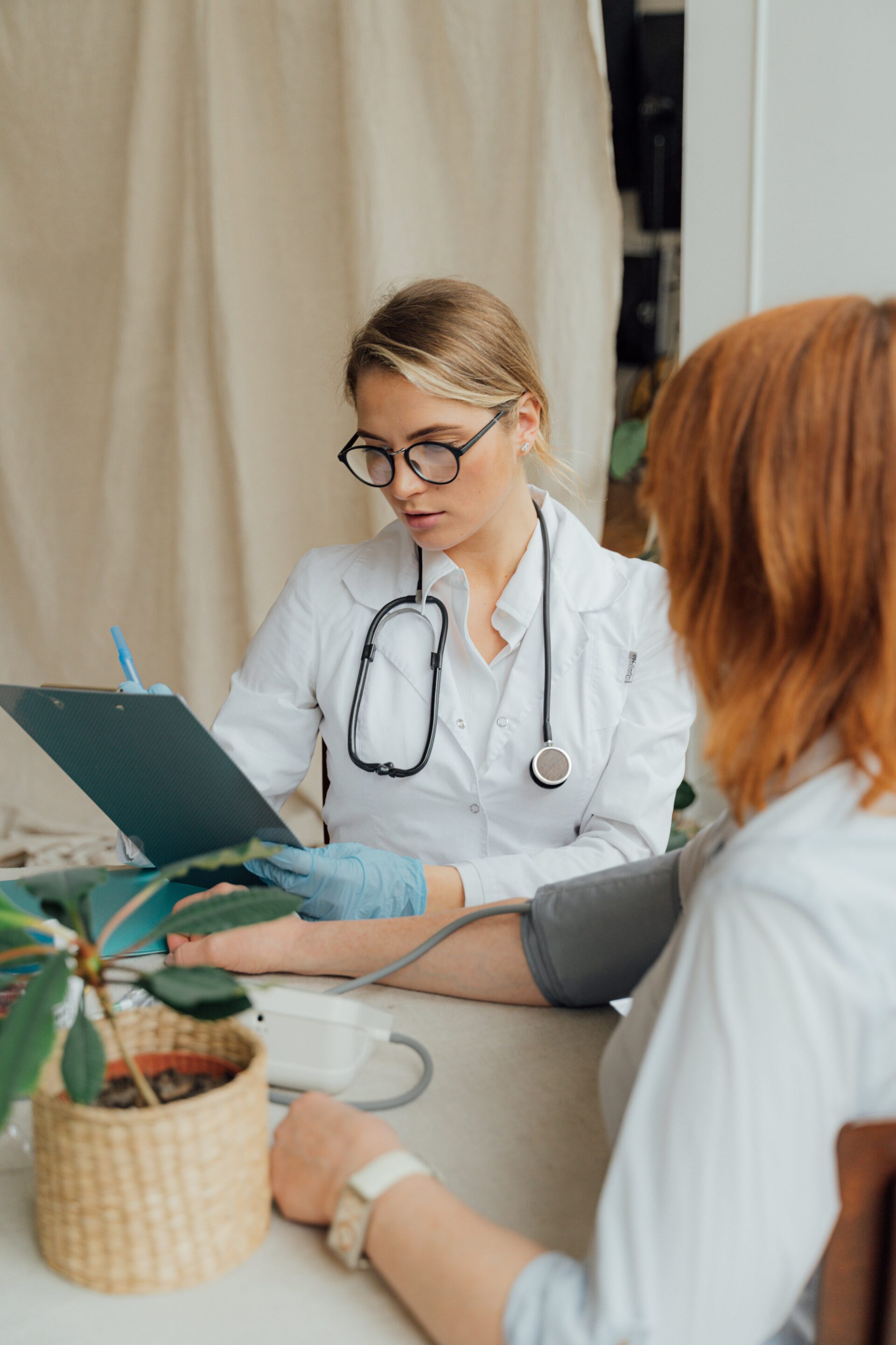 A female healthcare professional in a white coat and glasses is taking a patient's blood pressure. The patient, seen from the back, sits at a table with a potted plant in a woven basket. The medical professional is focused on her task, holding a clipboard in one hand and the blood pressure cuff in the other. They are in a room with a neutral-toned background.