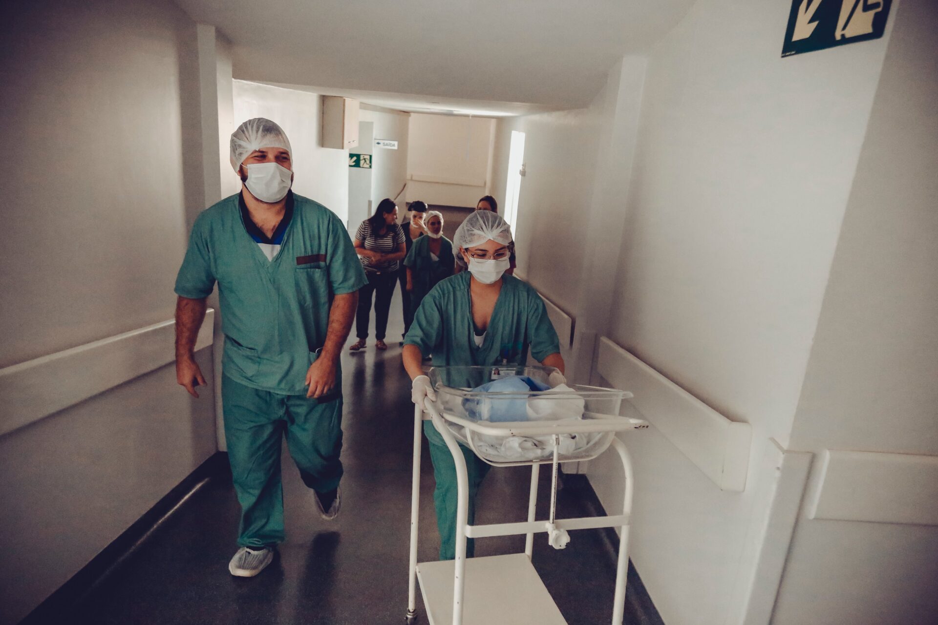 Two healthcare professionals in scrubs walking through a hospital corridor, one pushing a medical cart.