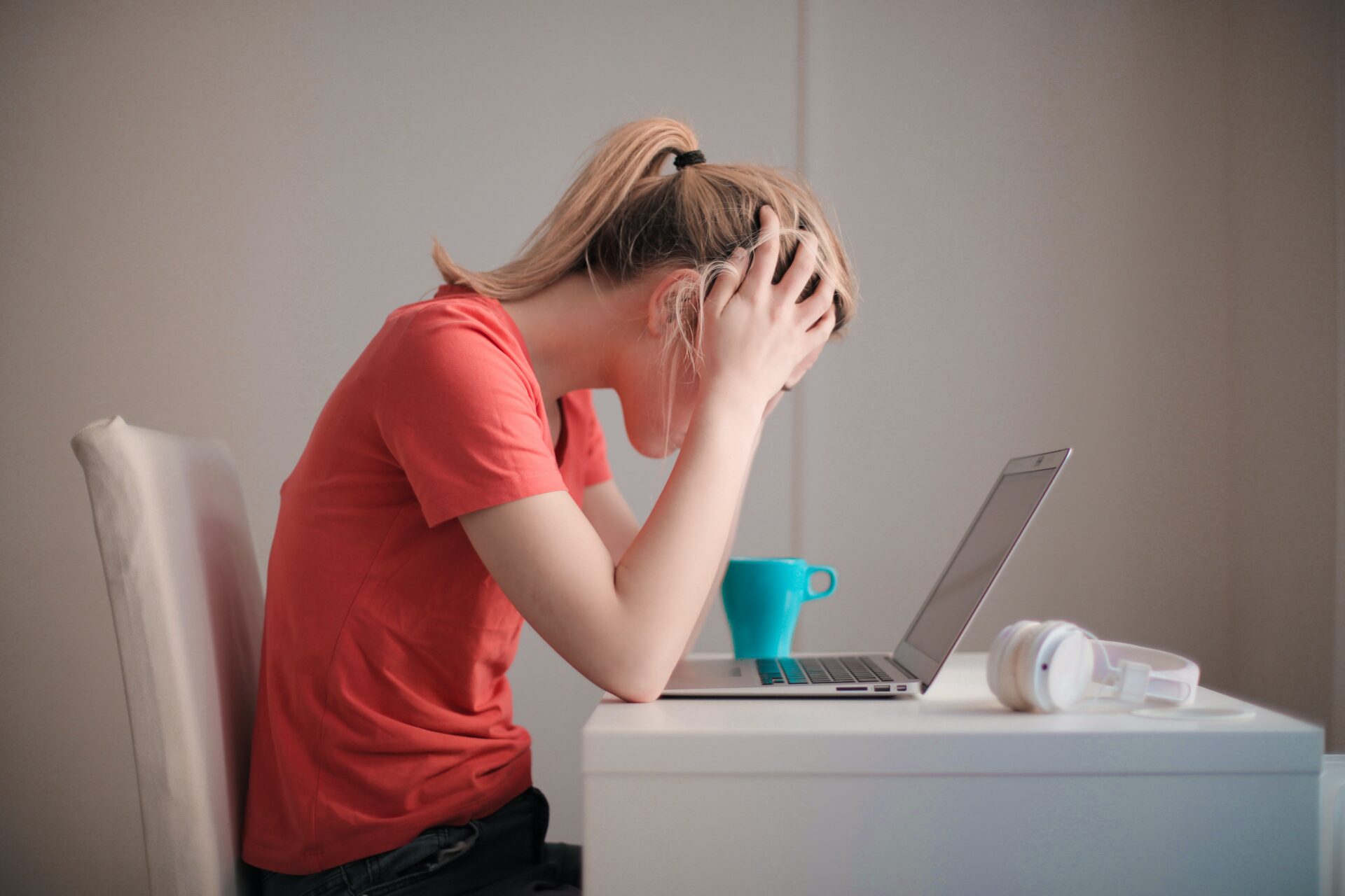 Stressed young woman with a ponytail facing laptop and holding head in hands, showing frustration or fatigue, with a blue mug and white headphones on the desk.