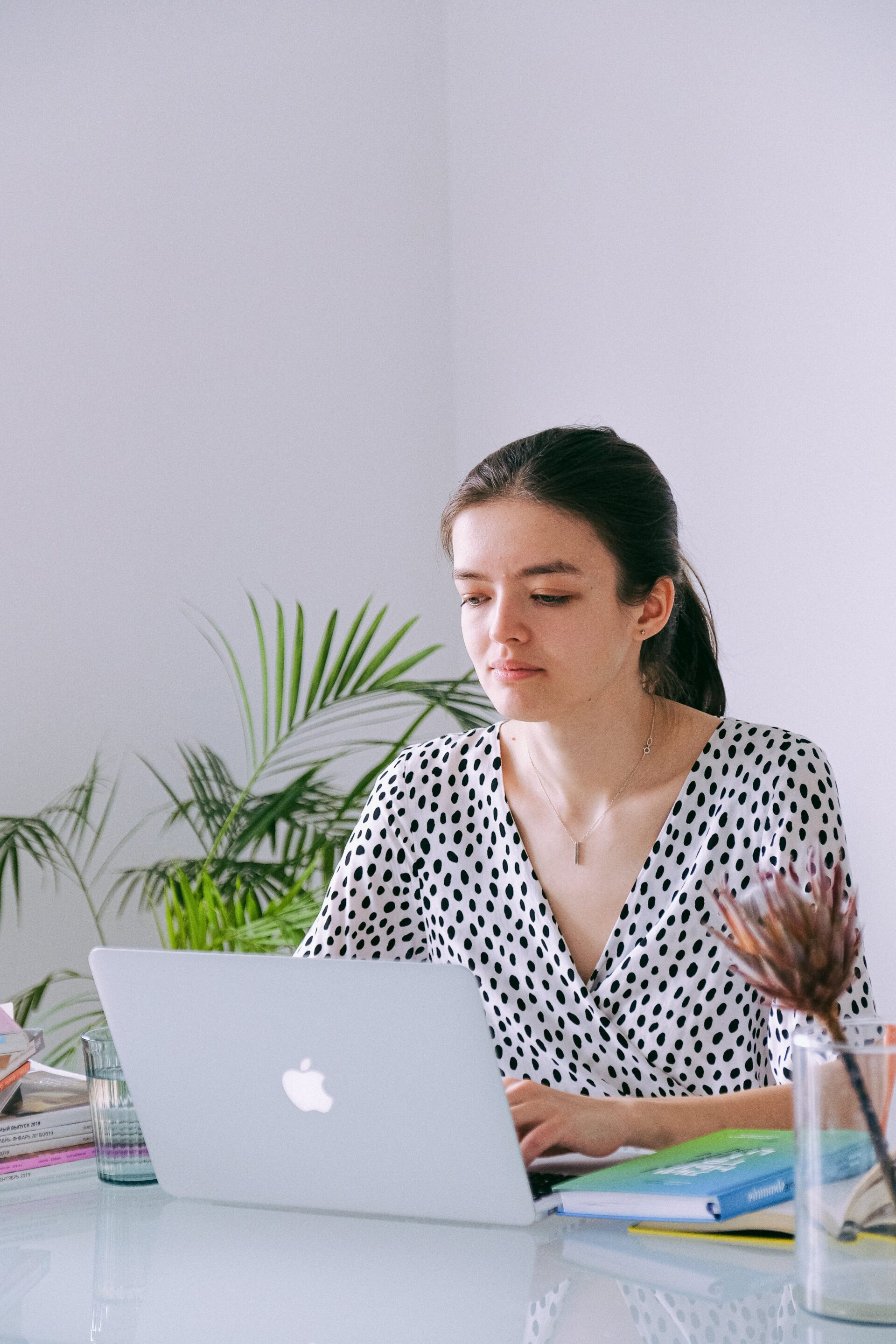 A focused woman with a polka-dotted top working on a MacBook at a clean and organized desk, with potted plants in the background, embodying a professional home office setting.