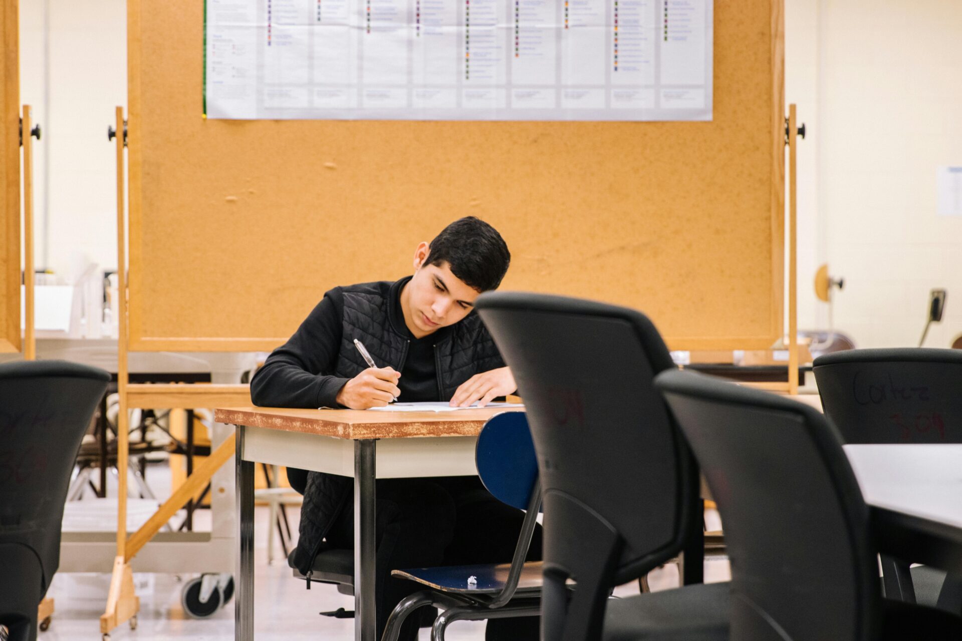 A young man focused on writing at a desk in a classroom, with chairs and a schedule board in the background.