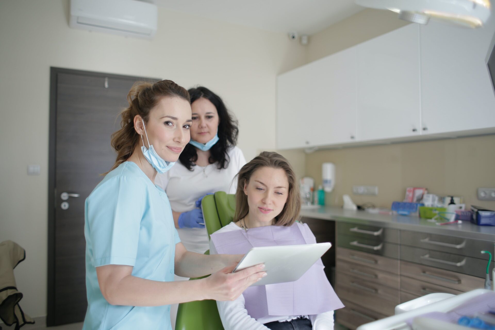 Dental nurse consulting with a patient in a clinic while holding a tablet, with a colleague standing by.