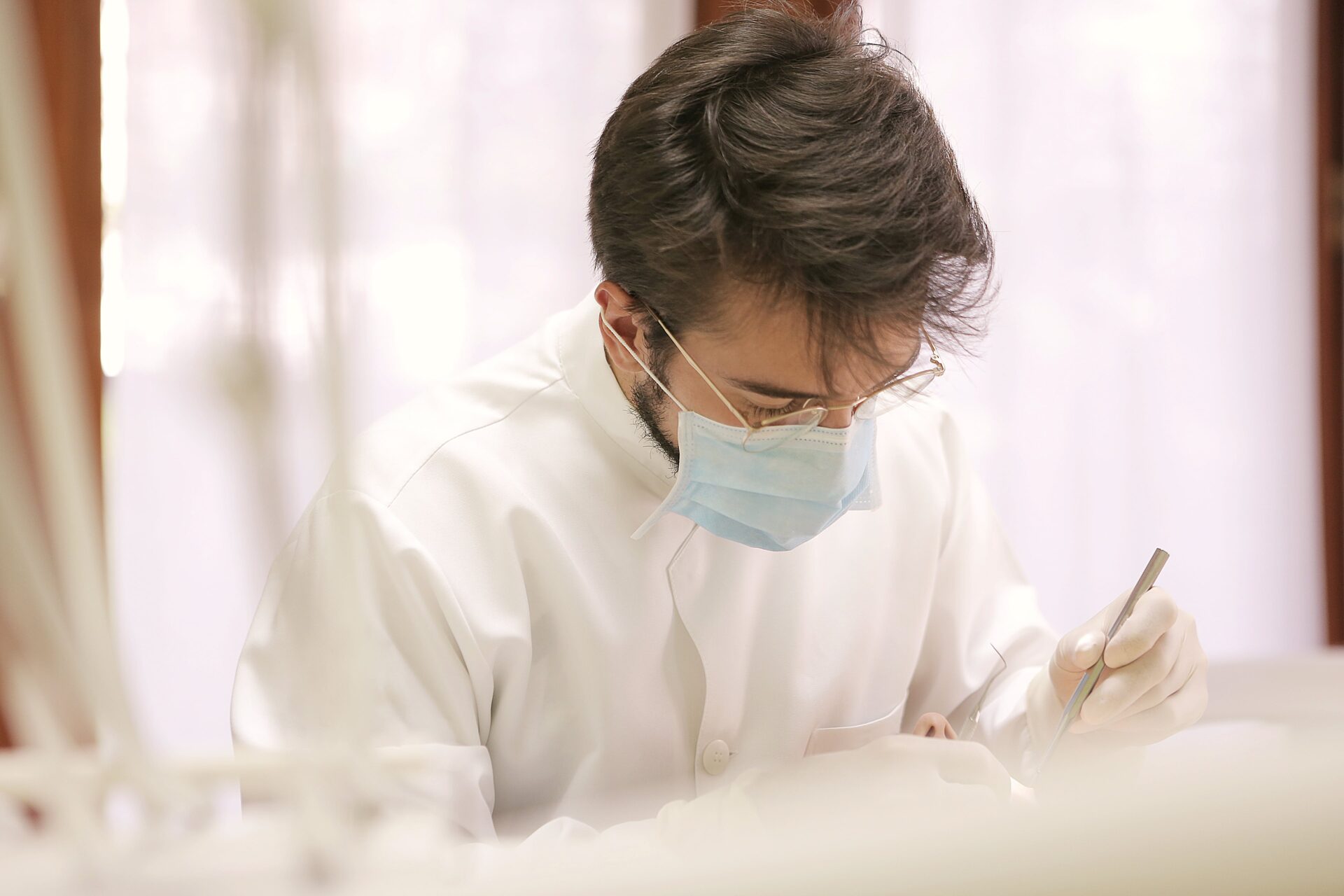 A focused healthcare professional wearing a mask and glasses is diligently writing on a clipboard, possibly updating patient records or completing medical documentation, in a brightly lit room.