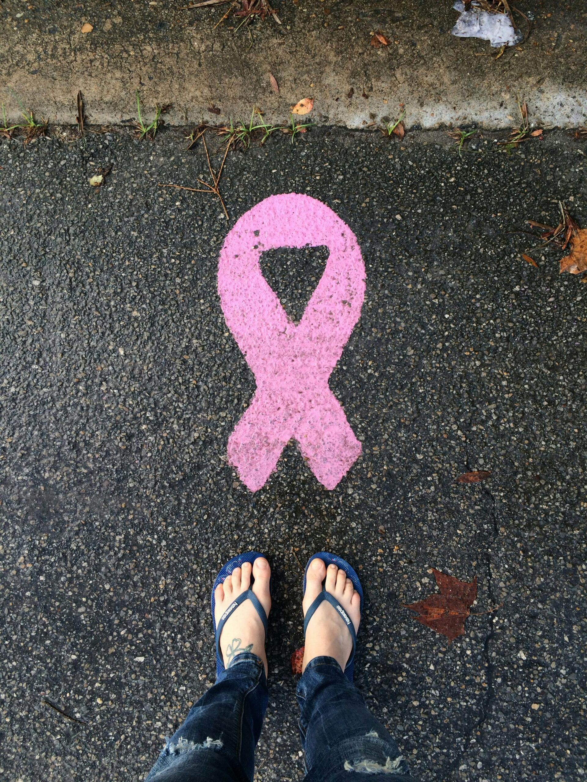 A pair of feet in blue flip-flops standing beside a pink ribbon painted on a wet asphalt surface, symbolizing support for cancer awareness.