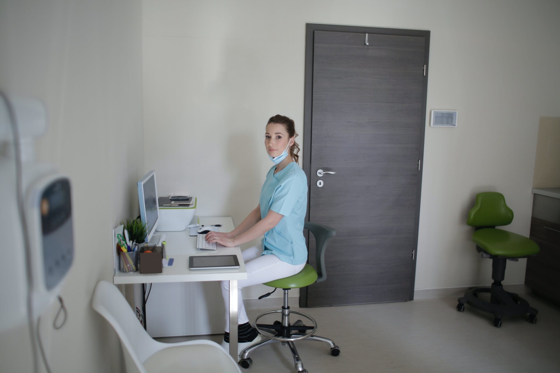 Nurse in scrubs sitting at a desk in a hospital office, nursing admission essay. The nurse is working on a computer with medical equipment in the background.