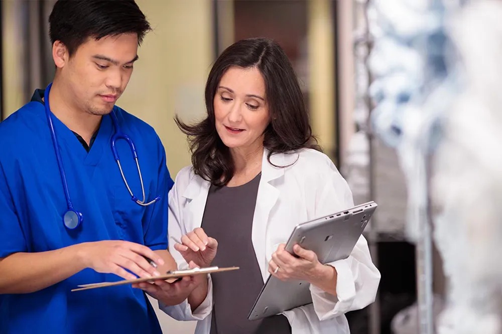 Two healthcare professionals, with their faces obscured for privacy, are engaged in a discussion. The individual on the left is wearing blue scrubs and has a stethoscope around their neck, holding a clipboard and writing. The individual on the right is wearing a white lab coat and holding a tablet computer. They appear to be in a medical setting, indicated by the blurred background which suggests clinical surroundings.