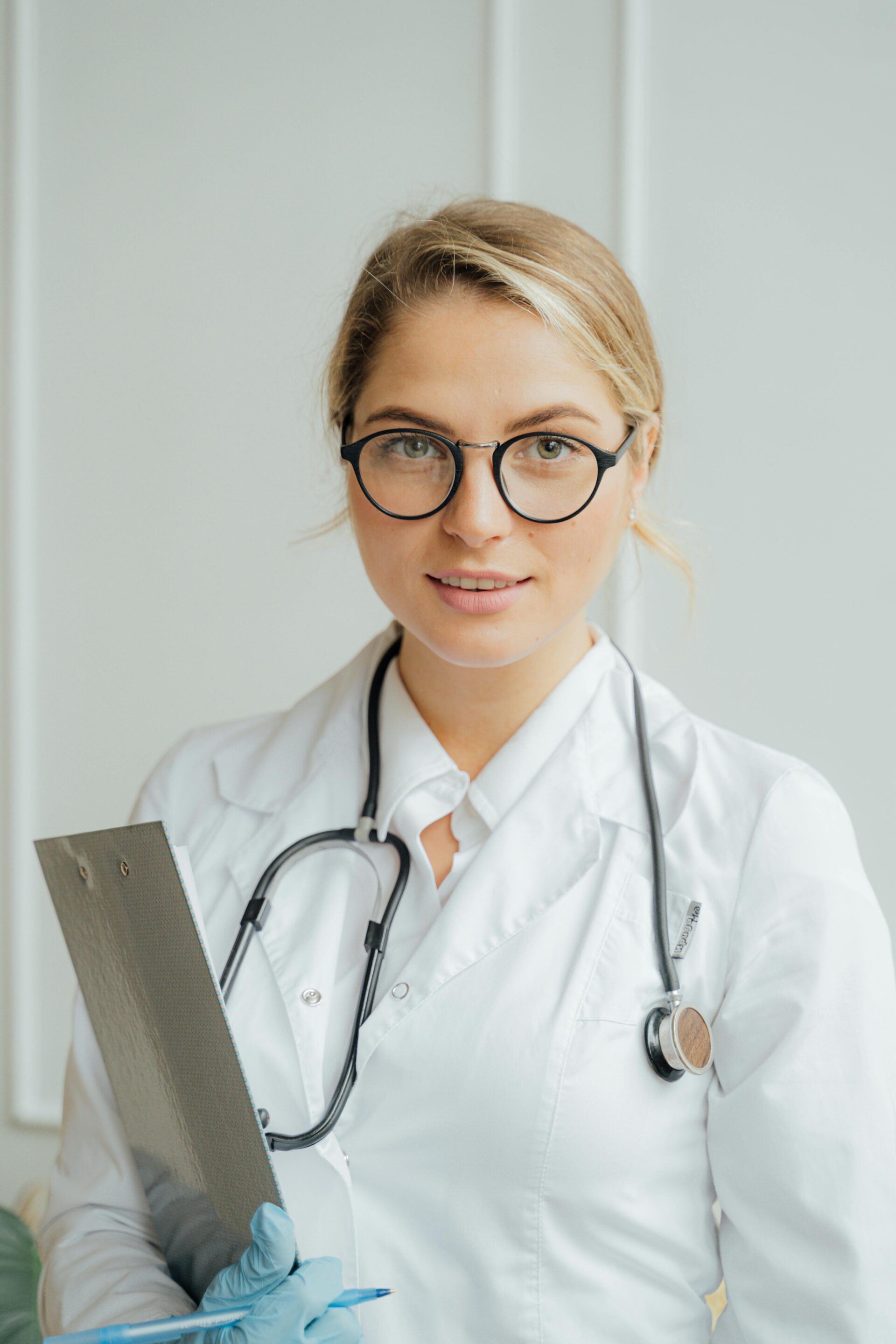 A female healthcare professional in a white lab coat and glasses, holding a clipboard with a nursing essay example and wearing a stethoscope around her neck.